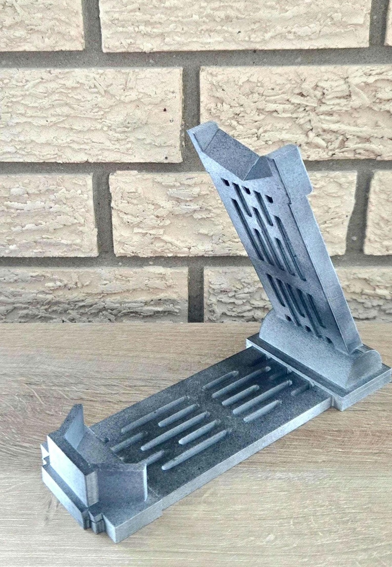 Op de afbeelding: Een grijze 3D-geprinte telefoonstandaard met een geometrisch ontwerp. De standaard is open en klaar om een telefoon te houden.