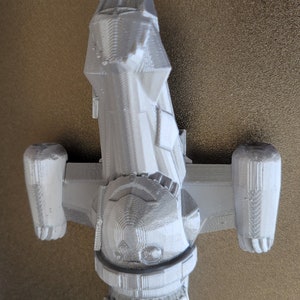 May include: A light gray 3D-printed model of a spaceship, featuring a central body with detailed panels, two side pods, and a rear engine. The model is set against a neutral background, showcasing the intricate design of the spacecraft.