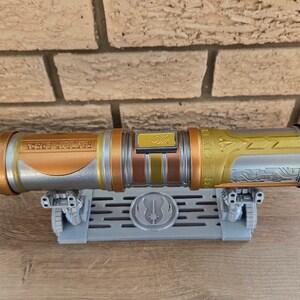 May include: A silver and gold lightsaber with intricate details and a black inscription on the hilt. The lightsaber is on a gray stand with a Jedi symbol.