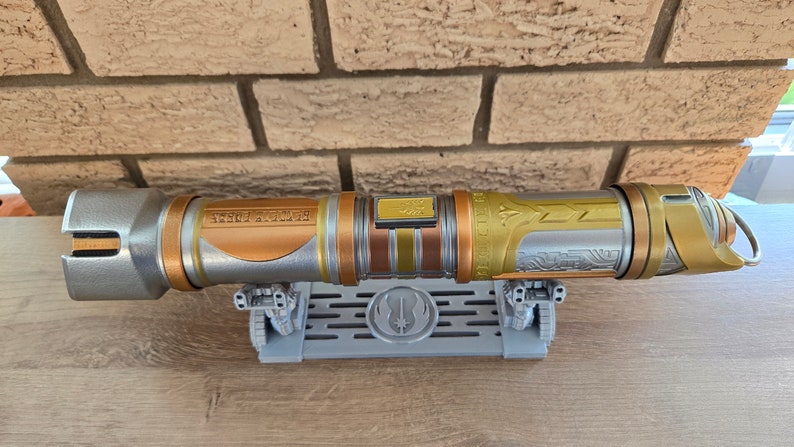 May include: A silver and gold lightsaber with intricate details and a black inscription on the hilt. The lightsaber is on a gray stand with a Jedi symbol.