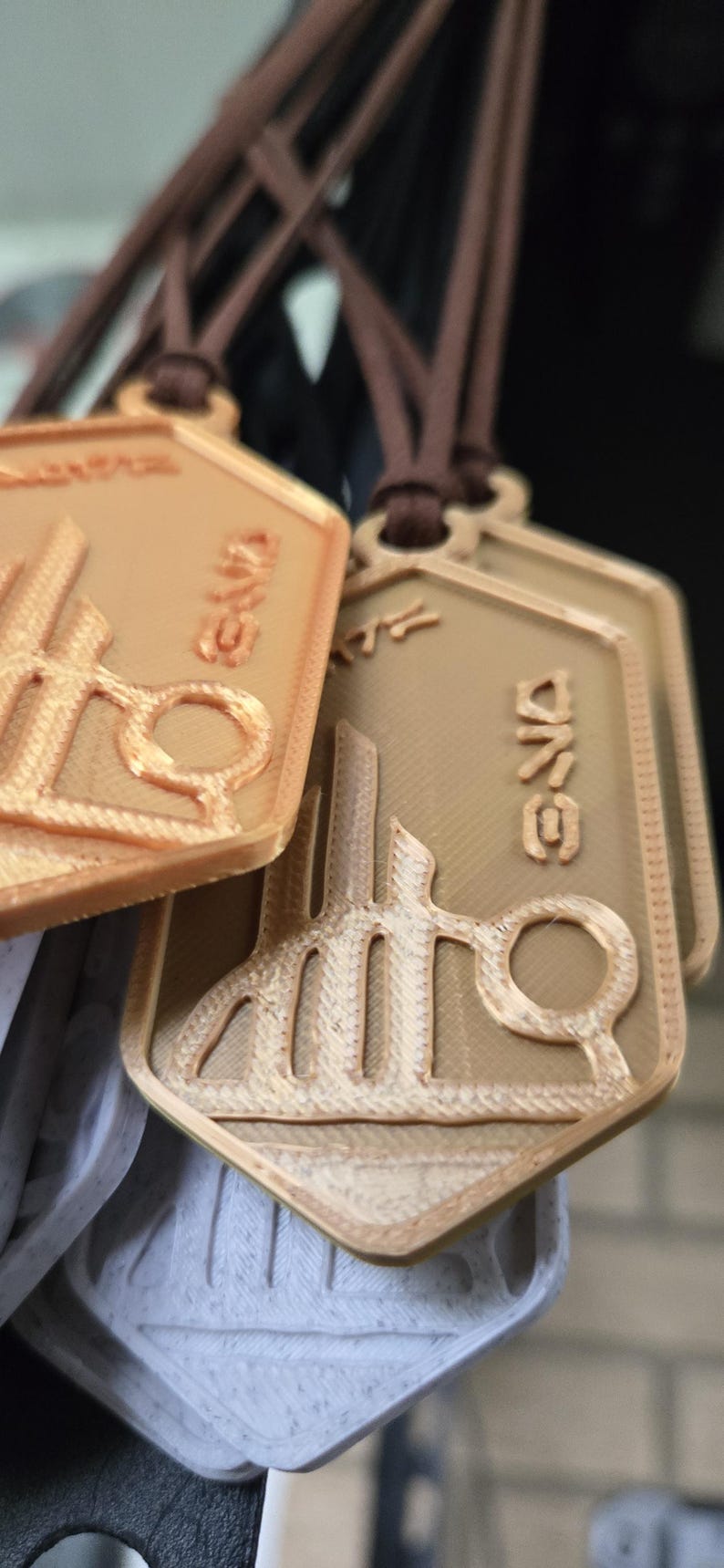 Puede incluir: Primer plano de varios colgantes de color bronce con un dise&ntilde;o de edificio y tijeras. Cada colgante es rectangular con esquinas redondeadas y cuelga de un cord&oacute;n marr&oacute;n. La palabra "GND" es visible en cada colgante.
