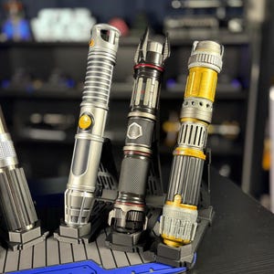May include: A collection of lightsabers in various designs, displayed on a black stand. The lightsabers feature silver, black, and gold accents. The hilts have intricate details, and the blades are not visible. The stand is blue and black.