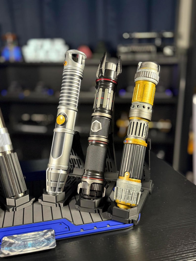 May include: A collection of lightsabers in various designs, displayed on a black stand. The lightsabers feature silver, black, and gold accents. The hilts have intricate details, and the blades are not visible. The stand is blue and black.