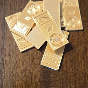 Może przedstawiać: Kolekcja złotych, prostokątnych element&oacute;w gry z wytłoczonymi wzorami. Niekt&oacute;re elementy mają okrągły emblemat, inne geometryczne kształty. Elementy są ułożone na ciemnej drewnianej powierzchni.