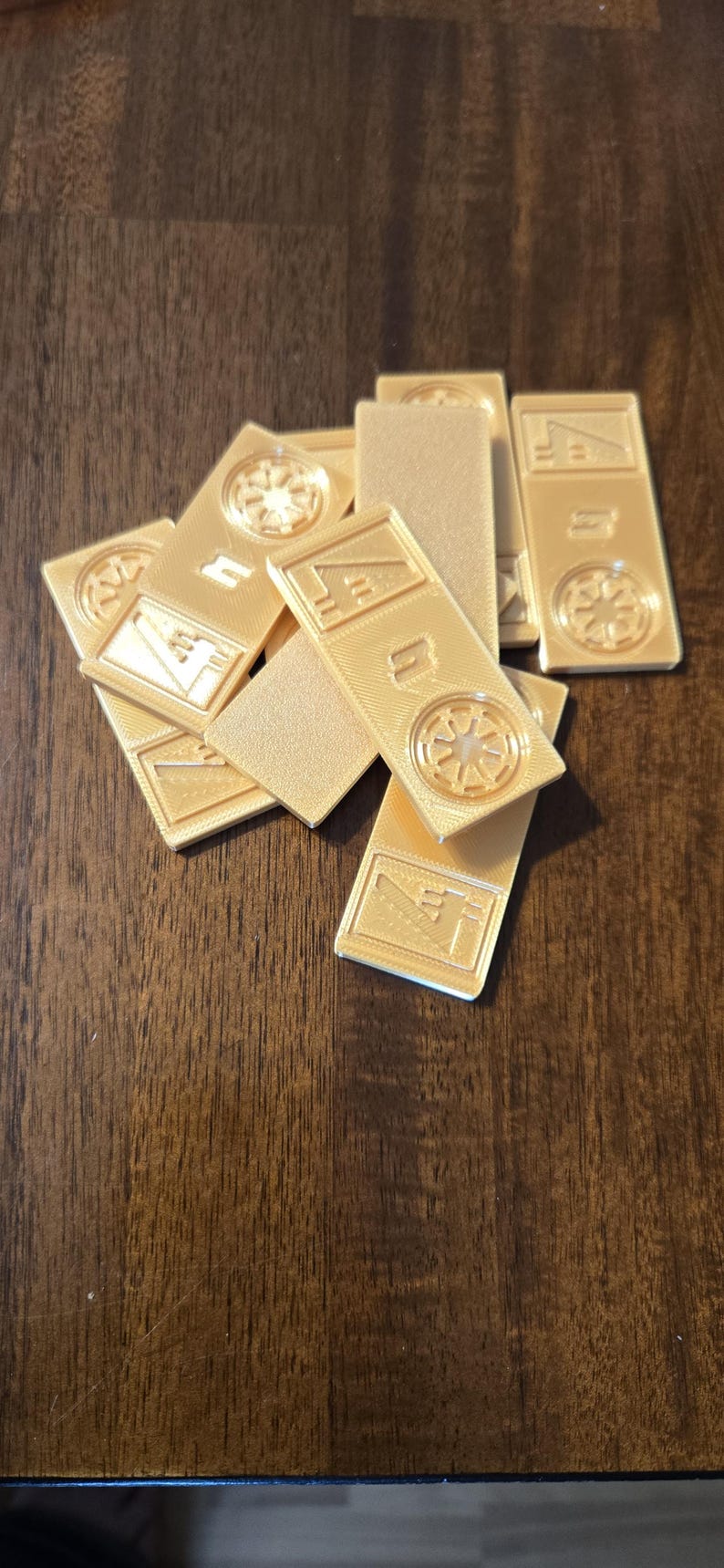 Może przedstawiać: Kolekcja złotych, prostokątnych element&oacute;w gry z wytłoczonymi wzorami. Niekt&oacute;re elementy mają okrągły emblemat, inne geometryczne kształty. Elementy są ułożone na ciemnej drewnianej powierzchni.