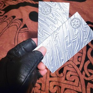 May include: Two silver metal plates featuring a raised design of the Star Wars Imperial symbol. The plates have a textured surface and are held in a gloved hand.