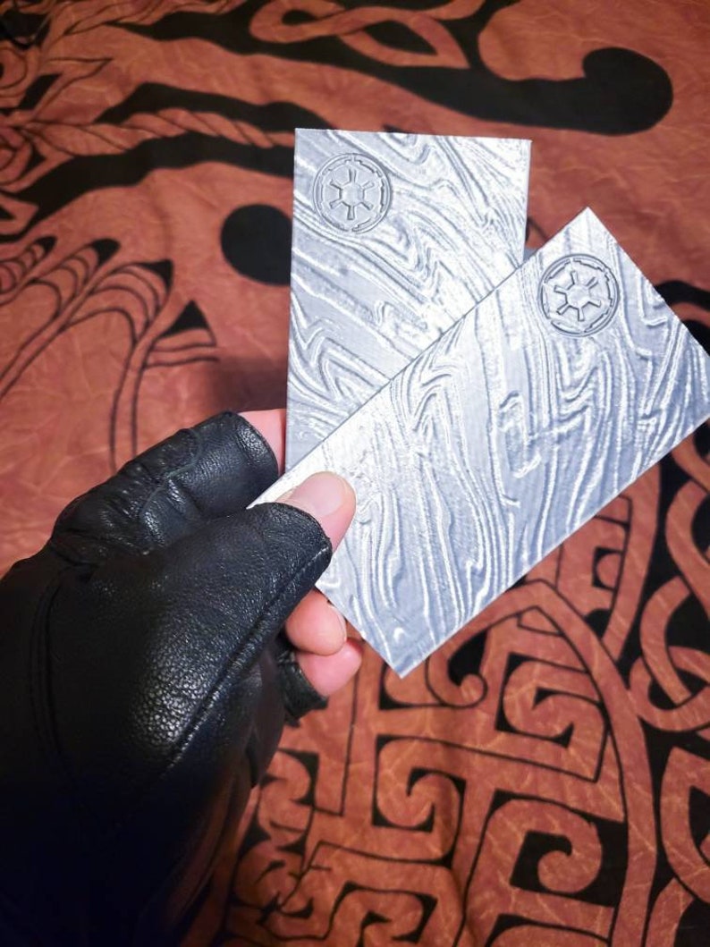 May include: Two silver metal plates featuring a raised design of the Star Wars Imperial symbol. The plates have a textured surface and are held in a gloved hand.