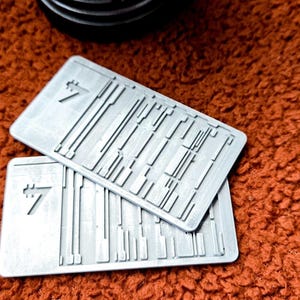 Peut inclure: Deux cartes rectangulaires en m&eacute;tal gris clair avec un motif textur&eacute; et le chiffre 7. Les cartes sont empil&eacute;es l'une sur l'autre. L'arri&egrave;re-plan est une surface textur&eacute;e brun-rouge.