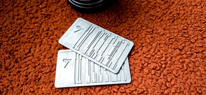 Peut inclure: Deux cartes rectangulaires en m&eacute;tal gris clair avec un motif textur&eacute; et le chiffre 7. Les cartes sont empil&eacute;es l'une sur l'autre. L'arri&egrave;re-plan est une surface textur&eacute;e brun-rouge.