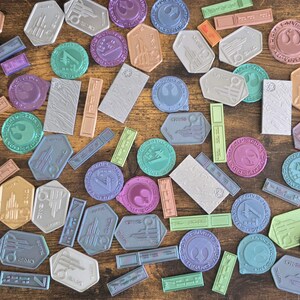 May include: A collection of colorful, round, and rectangular stickers with various designs. The stickers feature a variety of colors, including blue, green, purple, pink, and brown. The stickers are arranged on a wooden surface.