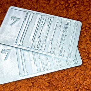 Peut inclure: Deux cartes rectangulaires gris clair avec une surface textur&eacute;e et un chiffre sept stylis&eacute;. Les cartes comportent une s&eacute;rie de lignes parall&egrave;les et perpendiculaires. Les cartes sont empil&eacute;es l'une sur l'autre.