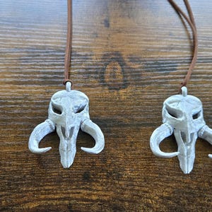 May include: Four identical pendants shaped like a stylized creature skull with curved horns, finished in a weathered silver-gray color. Each pendant is suspended from a thin, brown cord, creating a set of necklaces with a distinct, textured appearance.