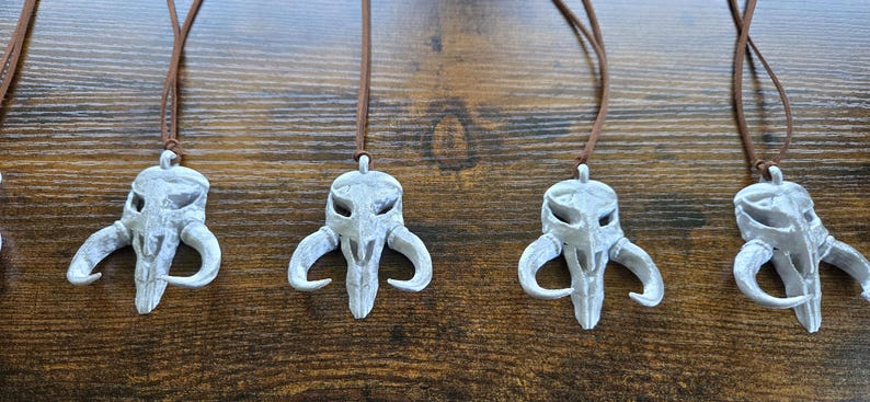 May include: Four identical pendants shaped like a stylized creature skull with curved horns, finished in a weathered silver-gray color. Each pendant is suspended from a thin, brown cord, creating a set of necklaces with a distinct, textured appearance.
