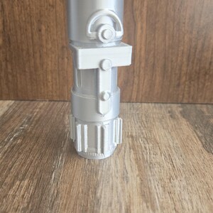 May include: A silver lightsaber hilt with a textured surface and a small, round button on the side.