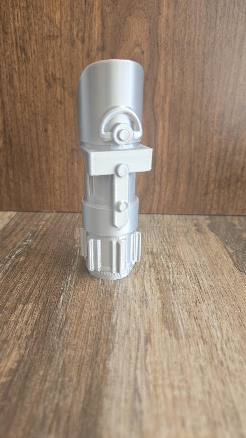 May include: A silver lightsaber hilt with a textured surface and a small, round button on the side.