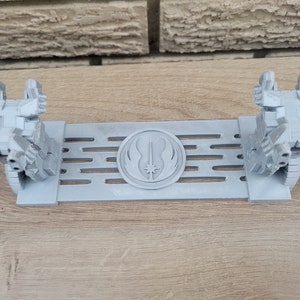 May include: A gray 3D printed stand with two figures on either side. The stand features a raised circular design with a lightsaber and the Jedi symbol in the center.