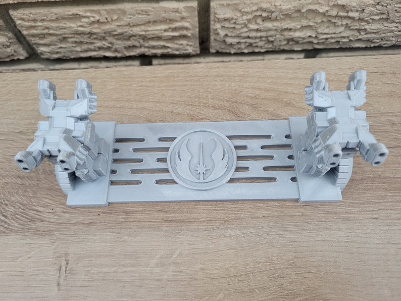 May include: A gray 3D printed stand with two figures on either side. The stand features a raised circular design with a lightsaber and the Jedi symbol in the center.