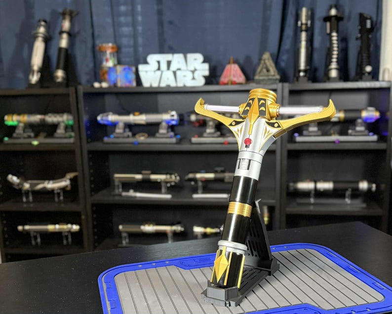 K&ouml;nnte beinhalten: Ein gold-, silber- und schwarzes Lichtschwert wird auf einem St&auml;nder pr&auml;sentiert. Weitere Lichtschwerter sind auf Regalen im Hintergrund zu sehen. Das Star Wars-Logo ist ebenfalls sichtbar. Das Lichtschwert hat einen Kreuzschutz.