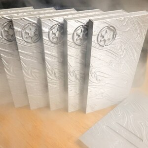 May include: A set of seven silver-coloured, rectangular, decorative blocks with a raised, embossed design. Each block features a stylized symbol of the Galactic Empire from Star Wars.