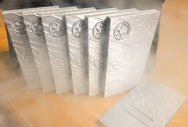 May include: A set of seven silver-coloured, rectangular, decorative blocks with a raised, embossed design. Each block features a stylized symbol of the Galactic Empire from Star Wars.