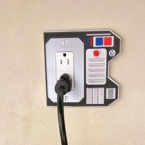 May include: A black and white wall plate with a standard electrical outlet and a decorative panel that resembles a control panel from a spaceship. The panel has red, blue, and white buttons and a red light.