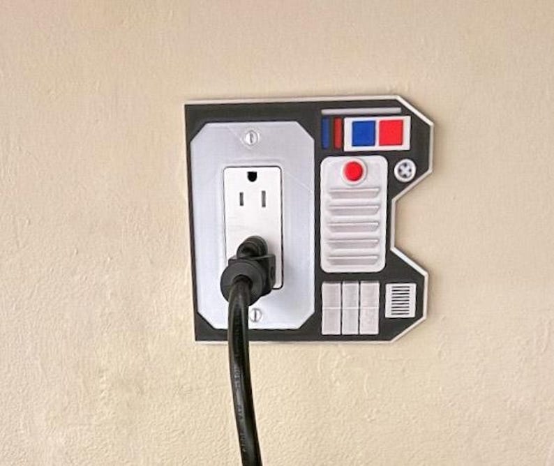 May include: A black and white wall plate with a standard electrical outlet and a decorative panel that resembles a control panel from a spaceship. The panel has red, blue, and white buttons and a red light.