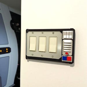 May include: A custom light switch plate with three light switches. The plate has a black frame with a silver center section. It features a control panel with red, blue, and silver accents, and is mounted on a white wall.