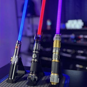 May include: Three illuminated lightsabers with blue, red, and purple blades are displayed on a dark surface. The hilts are detailed with metallic finishes and intricate designs, each resting on a black stand.