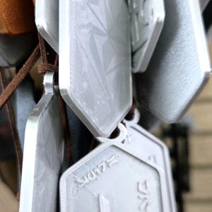 Puede incluir: Primer plano de varios colgantes plateados colgados de cordones marrones. Los colgantes tienen un dise&ntilde;o geom&eacute;trico y algunos tienen texto. Los colgantes probablemente forman parte de un collar o llavero.