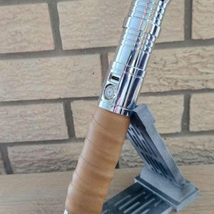 Op de afbeelding: Een zilveren en gouden lightsaber met een bruine leren grip, getoond op een grijze standaard.