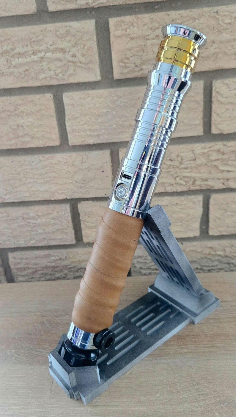 Op de afbeelding: Een zilveren en gouden lightsaber met een bruine leren grip, getoond op een grijze standaard.