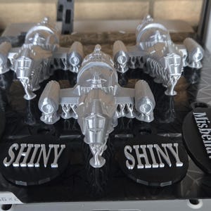 May include: A silver model spaceship with intricate details, displayed on a black base. The base features the word "SHINY" and the phrase "Aim to Misbehave" in white lettering. The overall theme is science fiction.