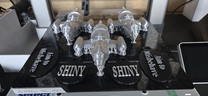 May include: A silver model spaceship with intricate details, displayed on a black base. The base features the word "SHINY" and the phrase "Aim to Misbehave" in white lettering. The overall theme is science fiction.