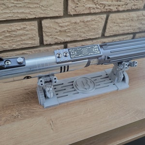 May include: A silver lightsaber with a black handle and a silver stand. The stand has a raised design of a symbol with a circle and a line through it. The lightsaber is on a wooden surface.