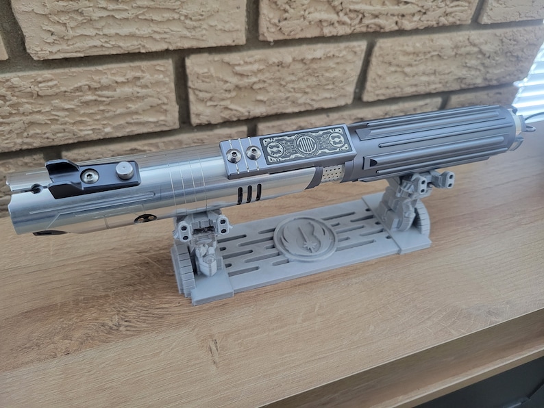 May include: A silver lightsaber with a black handle and a silver stand. The stand has a raised design of a symbol with a circle and a line through it. The lightsaber is on a wooden surface.