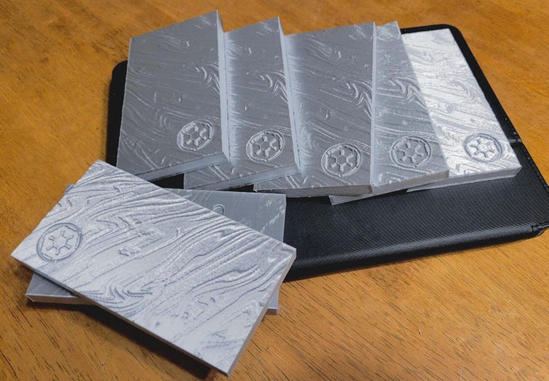 May include: A set of six silver-coloured rectangular tiles with a textured surface and a raised symbol. The tiles are stacked on top of each other and are sitting on a black leather surface.
