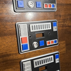 Op de afbeelding: Drie zwarte en zilveren lichtschakelaars met een Star Wars-thema. Elke schakelaar heeft een andere configuratie van knoppen en schakelaars. De knoppen zijn rood, blauw en grijs.