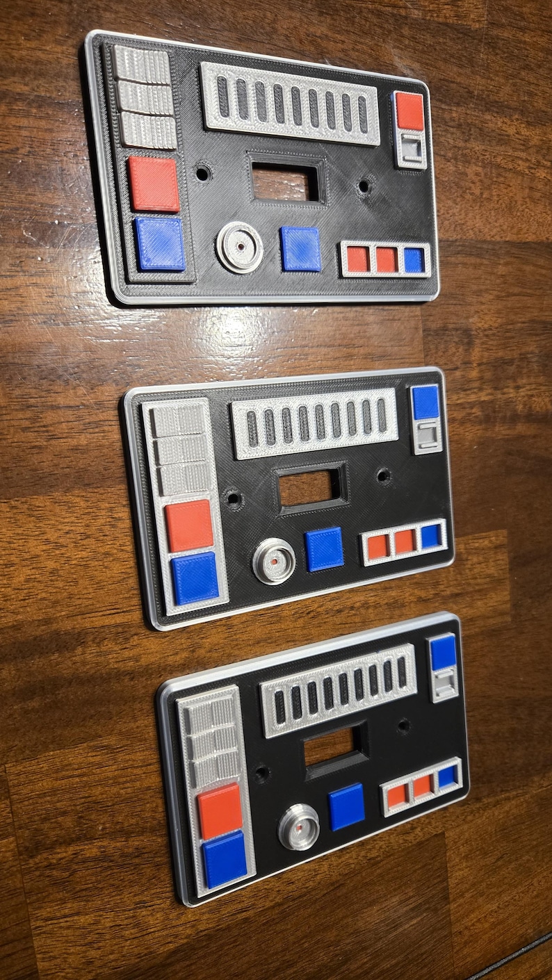 Op de afbeelding: Drie zwarte en zilveren lichtschakelaars met een Star Wars-thema. Elke schakelaar heeft een andere configuratie van knoppen en schakelaars. De knoppen zijn rood, blauw en grijs.