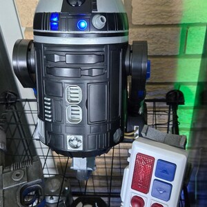 Peut inclure: Un jouet robot R2-D2 noir et argent&eacute; avec des lumi&egrave;res bleues, un panneau de commande blanc avec des boutons rouges et bleus, et d'autres jouets sur le th&egrave;me de Star Wars. Le robot a une t&ecirc;te cylindrique et un corps rectangulaire.