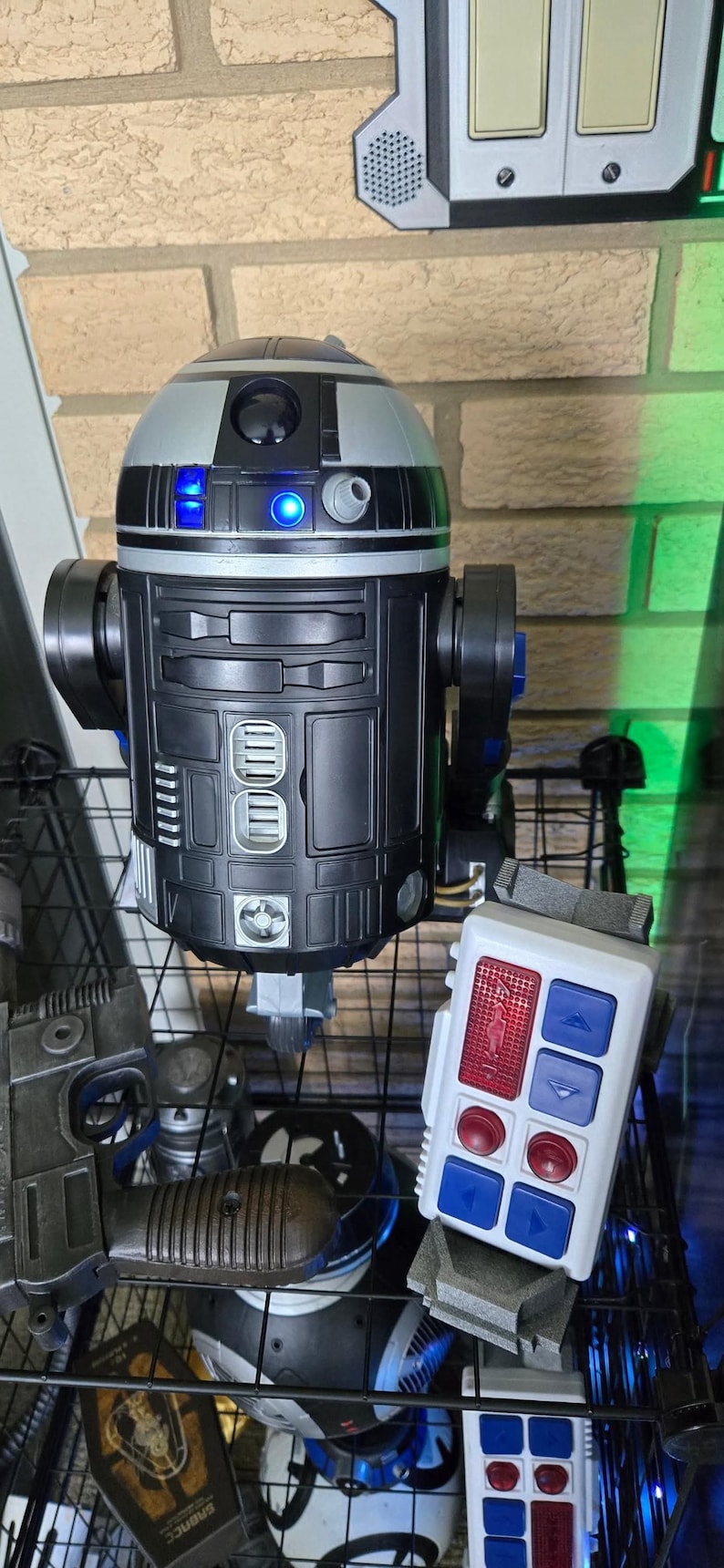 Peut inclure: Un jouet robot R2-D2 noir et argent&eacute; avec des lumi&egrave;res bleues, un panneau de commande blanc avec des boutons rouges et bleus, et d'autres jouets sur le th&egrave;me de Star Wars. Le robot a une t&ecirc;te cylindrique et un corps rectangulaire.