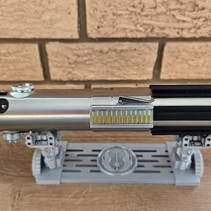 May include: A silver lightsaber with a black handle and a gold-colored emitter. The lightsaber is on a gray stand with a Star Wars logo.