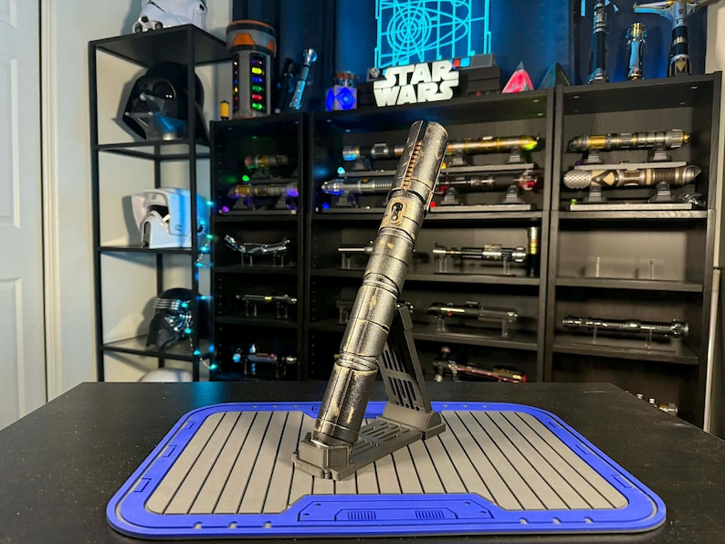 K&ouml;nnte beinhalten: Ein silbernes Lichtschwert auf einem schwarzen St&auml;nder, pr&auml;sentiert auf einer blau-grauen Matte. Das Lichtschwert ist der Mittelpunkt, mit einem detaillierten metallischen Design. Regale im Hintergrund zeigen verschiedene Lichtschwerter und Star Wars-Erinnerungsst&uuml;cke.