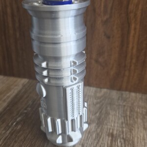 May include: A silver lightsaber-style lighter with a silver lighter inside. The lighter has a detailed design with grooves and ridges.