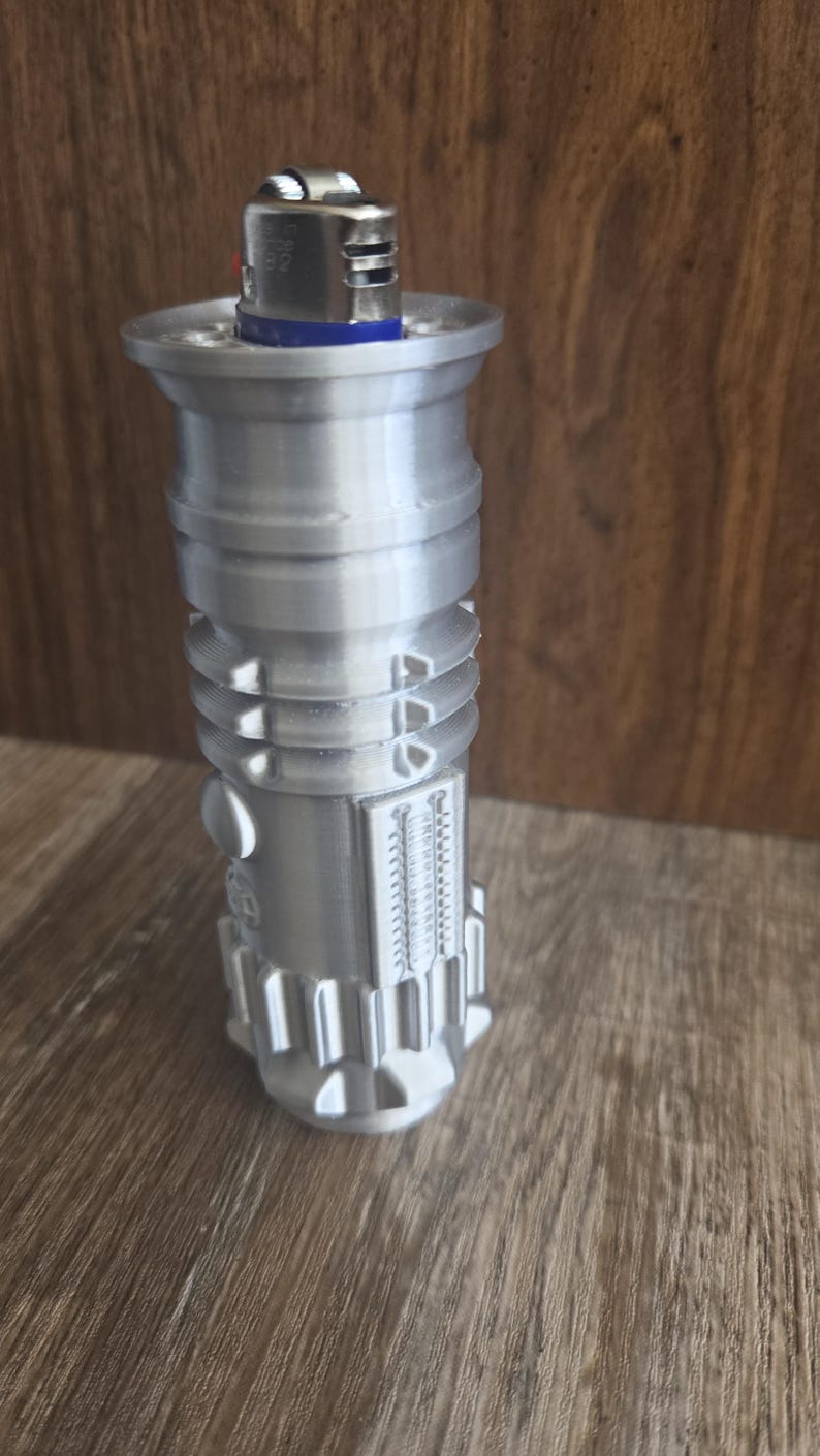 May include: A silver lightsaber-style lighter with a silver lighter inside. The lighter has a detailed design with grooves and ridges.