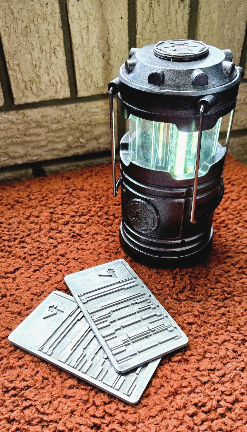 Peut inclure: Une lanterne noire &agrave; piles avec un panneau lumineux vertical transparent. La lanterne a une poign&eacute;e et repose sur une surface marron. Deux plaques rectangulaires grises avec un motif en relief sont devant la lanterne.