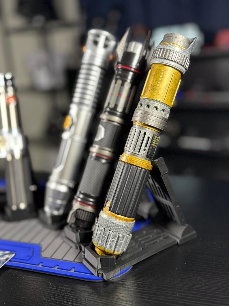 May include: A collection of three lightsabers, each with a unique design. The lightsabers are silver, black, and gold, and are displayed on a black and blue stand. The lightsabers are detailed with various textures and accents.