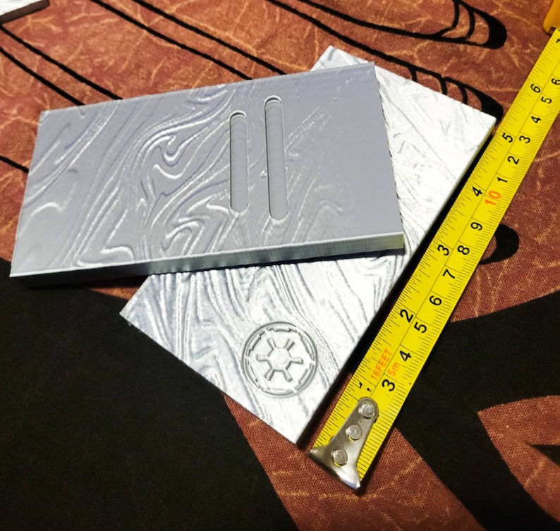 May include: Two silver-coloured plastic rectangular plates with a textured surface. The top plate has two rectangular cutouts. The bottom plate has a raised circular design with a star in the centre. A yellow measuring tape is partially visible in the bottom right corner.