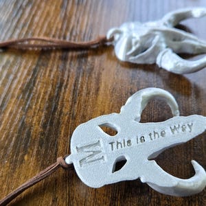 May include: Two white, matte-finished pendants shaped like stylized animal skulls, each attached to a thin brown leather cord. One pendant features the letter M and the text This is the Way embossed on its surface. The design is symmetrical.