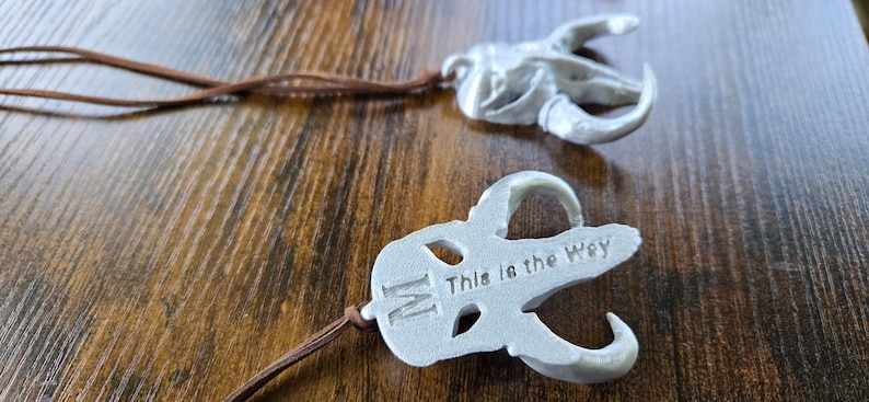 May include: Two white, matte-finished pendants shaped like stylized animal skulls, each attached to a thin brown leather cord. One pendant features the letter M and the text This is the Way embossed on its surface. The design is symmetrical.