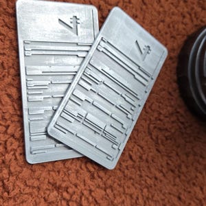 Peut inclure: Deux objets rectangulaires gris avec une surface textur&eacute;e et un symbole stylis&eacute;. Les objets pr&eacute;sentent une s&eacute;rie de lignes horizontales et verticales. Les objets sont sur une surface marron textur&eacute;e.